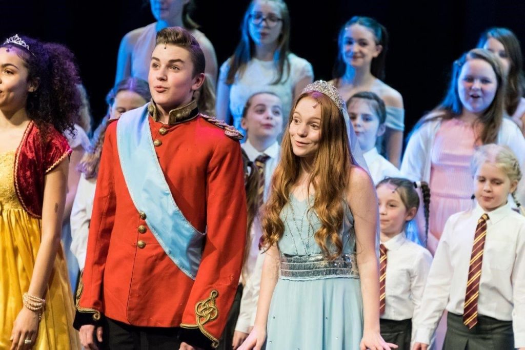 King's InterHigh pupil & musical actor, Daniel Wilmott, wearing a red military blazer & blue sash on stage, singing with fellow actors.