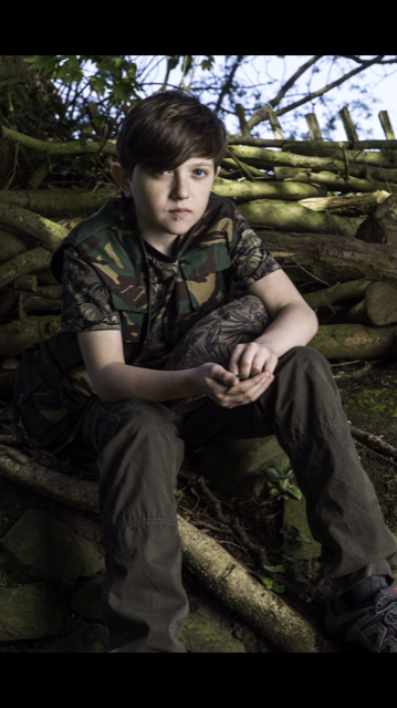 King's InterHigh pupil & actor, Gene Gurie, wearing army camouflage whilst sitting in front of a barrier made of sticks.